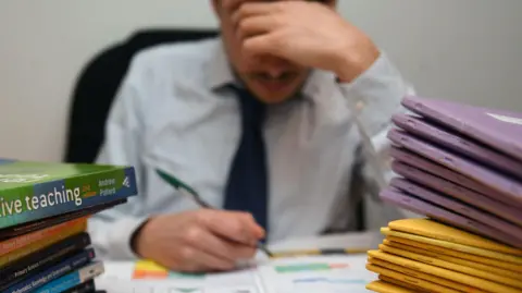 PA Media Photo of a school teacher looking stressed next to piles of classroom books.