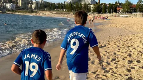 Family Photo The backs of a young Mackenzie and his younger brother wearing blue Everton shirts with the names of players Dominic Calvert-Lewin and James Rodriguez while walking on a sandy beach in Australia.
