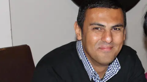 Akeela Mohammed A man with short dark hair and brown eyes is sitting at a table. He is wearing a blue and white checked shirt underneath a black jumper.