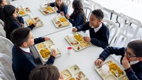 Getty Images Children in school uniform eat school dinners from trays and are laughing and talking together