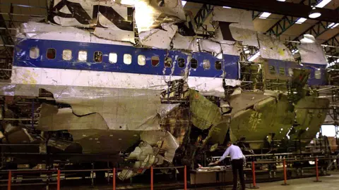 Reuters The wreckage of the plane in a warehouse. It is white with a blue stripe and black lettering, and it has missing and damaged sections. A man in a white shirt and black trousers is standing in front of the wreckage.