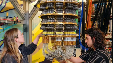 Google Google staff, a woman to the left and a man to the right work on the cryostat which holds the chip and keeps it very cold. The cryostat losley resembles a chandelier made of cascading thin metal tubes.