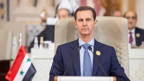 Reuters Basar al-Assad attends Organisation of Islamic Cooperation (OIC) summit in Riyadh, Saudi Arabia, November, 11, 2023 - he sits slightly slumped in his seat with a serious expression on his face, and the Syrian flag visible in the near ground. He is wearing a blue suit and blue tie.