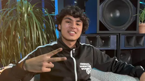 Haydog sits in a white cushioned chair. He holds up a V sign with his right hand. He's wearing a zipped up black shell top and grey trousers. There's a plant and a big speaker in the background.