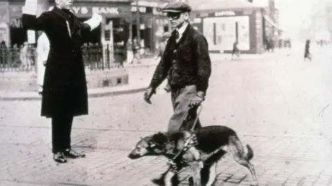 Guide Dogs A black and white image of a man kneeling in a field with a Labrador dog. The man is wearing a suit, checked shirt and striped tie. The dog is on a lead.