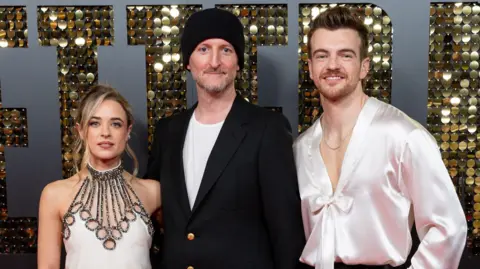 Getty Images Raechelle Banno is dressed in a white dress, Michael Gracey has a black blazer over a t-shirt and Jonno has a silk shirt. They are at a premiere event for Better Man.