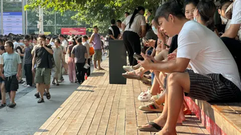 BBC/Xiqing Wang Crowds at a job market in Guangzhou city - people sit in rows on steps, while a crowd walks past.
