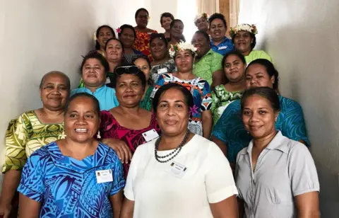 UNDP Dr Puakena Boreham, centre, wearing white, surrounded by 20 women, all smiling, some in traditional Tuvaluan dress with flowers in their hair, in a photo from 2018
