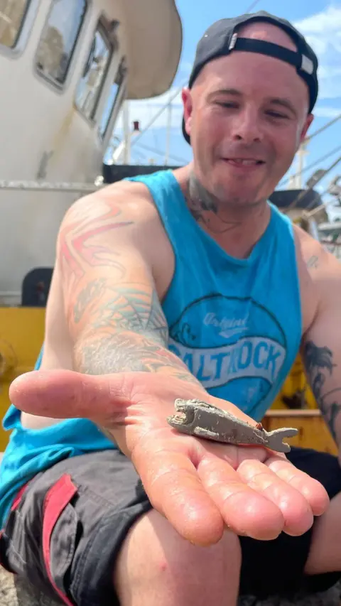 Richard West Richard West is sitting on a trawler and has stretched his open palm out towards the camera. In his hand is a pale grey barnacle-covered Lego shark and he is smiling. He has tattoos on his arms and is wearing a blue cutaway Saltrock T-shirt and a backwards cap.