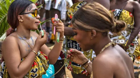 Olympia De Maismont / AFP A woman in silver carnival skirt and waistcoat outfit helps a fellow participant in matching clothes lace up her gold boots - Calabar, Nigeria