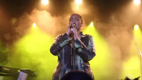 Kingpin @djkingpin33 A woman on stage, singing into a microphone, with yellow lights and smoke in the background. The singer is holding the microphone with both hands and wearing a black and grey dress.