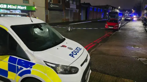 BBC A street with a police cordon. The sky is dark and street lights are on. There is a police car at the front of the image, with a police cordon, a red car, and a blue bus behind it on the street. There is a corner shop with a green and yellow sign on the left of the street.