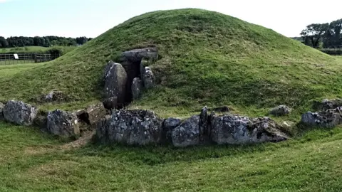 Prof George Nash A circular stone structure with upright pillars surmounted with a capstone covered in turf.