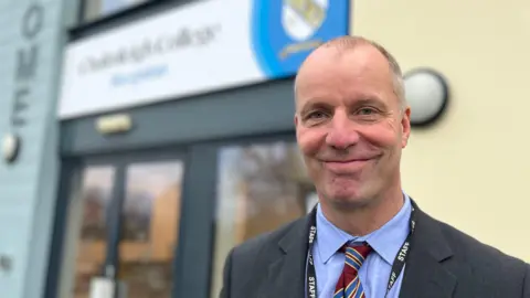 BBC Michael Johnson, head teacher at Chulmleigh College, wearing a blue shirt, a burgundy striped tie, grey suit and a staff lanyard in front of the school