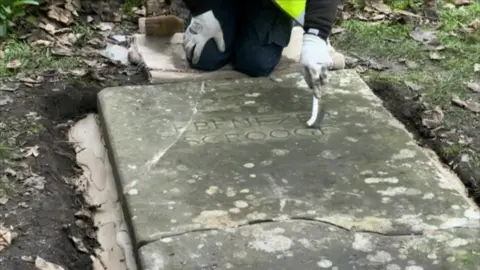 BBC A person is kneeling above a gravestone that is lying in the ground. The stone has cracks over it. The person is using a thin brush to clean inside letters that read "Ebenezer Scrooge"