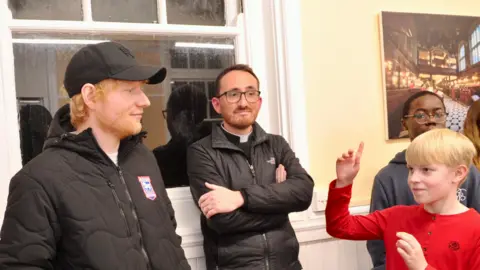 Lucy Taylor Photography Ed Sheeran wearing a black cap and black Ipswich Town jacket and looking towards a fair haired boy, wearing a red top, and with his right hand raised. Between them leaning against a window is a vicar wearing his dog collar and leaning against a window frame with his arms crossed.