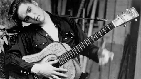 Michael Ochs Archives/Getty Images Elvis Presley poses for a portrait holding an acoustic guitar in 1956. He is wearing a dark-coloured shirt and is resting his head against a tree.