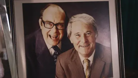 Hansons Eric Morecambe laughing at the camera with his signature, black framed glasses, with Ernie Wise to the right of him, smiling at the camera