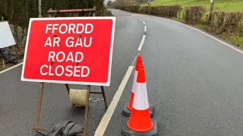 BBC A red sign on a road stating it is closed. To the right of the sign are two cones. The road is in a rural setting