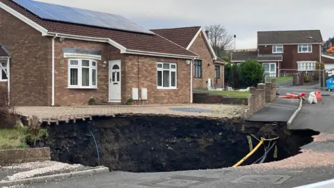 BBC Large sinkhole in street on the drive of one house and into the road, with other homes nearby, barriers in the street.