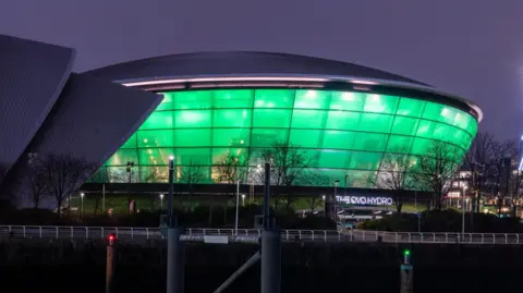 Getty Images A large events arena with its windows lit up in green in front of a river
