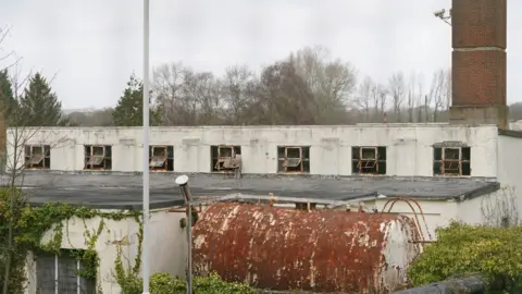 PA Media A derelict white building with a large, rusted metal container in front of it. To the right is a large red brick chimney