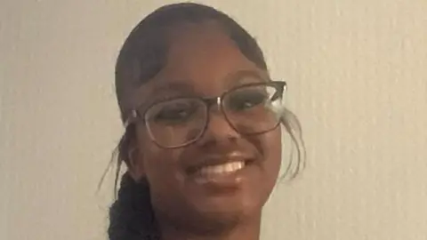 PA Media A close-up for a smiling Elianne Andam, she has long black hair in a braid and is wearing glasses. She is wearing a striped school uniform blouse.