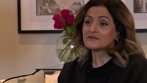 BBC A woman with dark hair and brown eyes sits on a sofa. She is wearing a black top and jacket and earrings. Behind her are two artworks on a wall and a vase of pink roses.