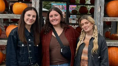 Casey Galt Three girls in their early 20s smile with pumpkins behind them on shelves.