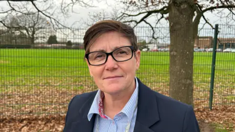 BBC A woman with short brown hair and black framed glasses in a blue jacket and blouse standing in front of a fence with a leaf-covered field behind it