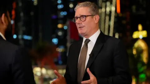 Reuters Sir Keir Starmer, who has a grey 'short back-and-sides' hairstyle and wears black-rimmed glasses, gestures with his hands as he speaks to the media after arriving in Abu Dhabi