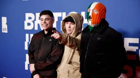 Getty Images Kneecap at 2024 BIFAs - left to right Móglaí Bap in black Lacoste zip jacket, Mo Chara in a cream jacket with hood up, wearing a black snapback and pointing at camera, and DJ Próvaí in black jacket and green white and orange tricolour balaclava