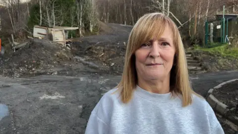 BBC Dianne Morgan, with shoulder length blonde hair and wearing a grey jumper, is standing in front of a coal tip. Behind her you can see the area of the coal tip which caused the landslide. There are remains of a shed which was badly damaged and there are also trees surrounding the coal tip area.