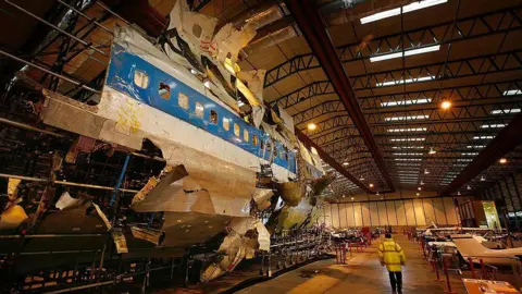 Getty Images The wreckage of the plane has been reconstructed in a warehouse. It is white with a blue stripe and black lettering, and it has missing and damaged sections. A person in a high viz jacket is walking next to the wreckage.