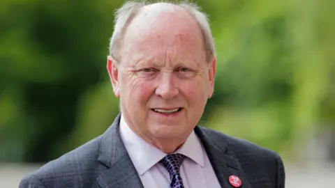 PA Media Jim Allister in a grey checked blazer smiles to camera. He has grey hair and is wearing a lilac shirt with purple spotted tie. On his right lapel he wears a badge reading 'no sea border'.