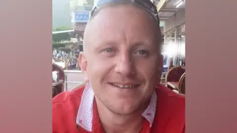 Family A man with cropped fair hair and wearing a red jumper and sunglasses on his head smiles at the camera. He is sitting on a restaurant terrace. 