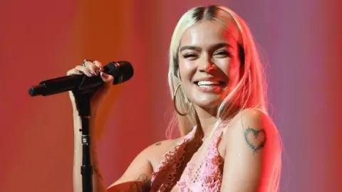Getty Images Colombian singer Karol G holds a microphone and smiles as she performs at a concert in Los Angeles