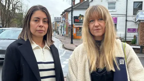 BBC Two women stand next to each other facing the camera, with a busy high street behind them. The woman on the left has dark shoulder length hair and is wearing a cream and blue striped top and a navy jacket, the woman on the right has long blonde hair and is wearing a cream fleece