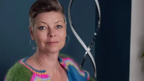 Sheila Foster-Hancock Laura Marfell-Williams, with swept back short hair and hooped earrings. She is wearing a striped green, bright pink, grey, blue and yellow woollen cardigan on top of a white vest top, and is sitting against an inky blue wall.