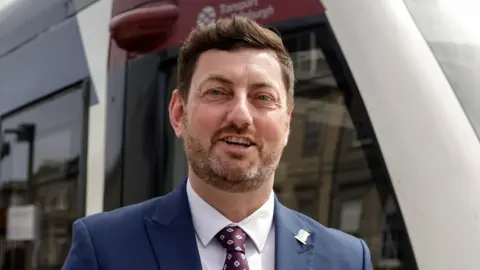 Getty Images Cammy Day is smiling at the camera. He is wearing a suit and standing infront of a tram.