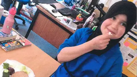 Surrey Police Sara Sharif wearing a black hijab and blue outfit. She appears to be in a classroom of office. She is holding a piece of food near to her mouth.