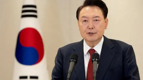 Reuters South Korean President Yoon Suk Yeol delivers an address to the nation at the Presidential Office in Seoul, South Korea. He wears a dark blazer, a red tie and stands to the side of the South Korean falg.