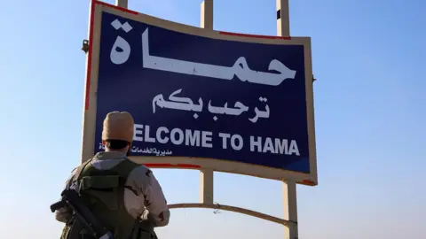EPA A rebel fighter stands next to a sign saying "Welcome to Hama" on the Damascus-Aleppo highway, Syria (3 December 2024)