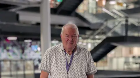 BBC Steve Mitchell, has silver short hair, and stands centre infront of the glass rail on the first floor inside BBC central square building in Cardiff. He wears a white and grey polo shirt and has a purple visitor lanyard around his neck. 