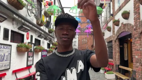 Beyond Skin/ Smack Jay The Zambian rapper, Smack Jay, standing in an alleyway in Belfast. To his left and right are the entrances to two pubs. Above him are coloured lanterns hung across the alleyway.   