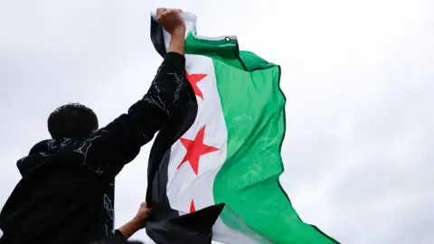 EPA A man in a sweatshirt, his back to the camera, waves a Syrian opposition flag, with stripes of green, black and white with red stars on it, in the air