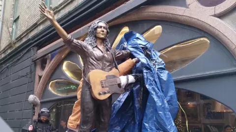 A bronze statue depicting the musician Rory Gallagher. He has his hands in the air, and a blue tarpaulin sheet is being pulled off. Behind the statue is a brick building.