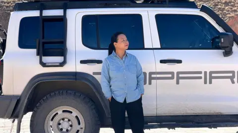 Su Min Su Min, wearing a blue shirt and black pants, standing in front of her white Volkswagen hatchback