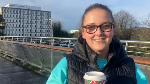 Cynthia is standing on a bridge and holding a disposable cup of coffee. She wears tortoiseshell glasses and has a black body warmer on over a green jumper. Marlborough House is in the background.