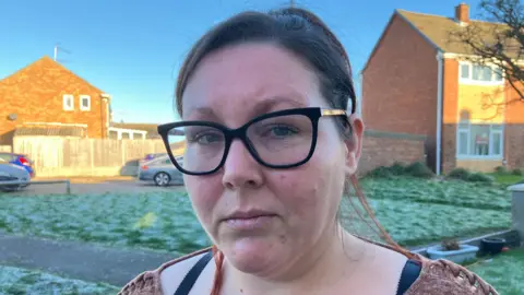 Sam Read / BBC A woman with black hair and wearing black rimmed glasses and a brown top standing in front of a grassy area with terraced houses in the background. 